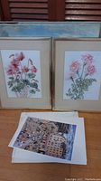 Two framed Kathleen England flower watercolours with pink flowers, beige matting, silver frames, and three cityscape prints stacked on the floor.