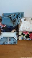 Photo showing stacked folded blankets in solid blue, floral prints, and cream color, along with a partially visible fabric bolster pillow.