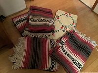 Four folded striped blankets in red, black, white, and gray with fringe and one cream needlepoint textile with geometric embroidery.