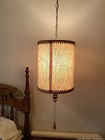 Photo of one vintage hanging lamp shade illuminated, showing textured cream fabric and scalloped trim with pull chain and tassel.
