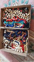 Two cardboard boxes containing assorted costume necklaces made of beads, shells, and wood in colors including red, blue, white, black, turquoise, and brown.