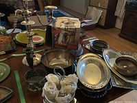 Overview of vintage silver plated trays, bowls, brass planters, candlesticks, and boxed items on wooden surface