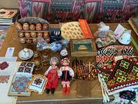 Overview photo showing all items including wooden eggs, dolls, needlepoint pieces, and textiles on table.