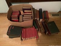 Photo of approximately 30 hardcover vintage books stacked in a cardboard box and arranged on the floor, showing various colors and bindings