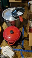 Overview photo showing all items including three cast iron fry pans, red Fontignac enameled stock pot, and aluminum stock pot in a clustered arrangement on carpeted floor