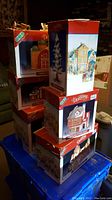 Stack of seven Lemax Dickens' Village Christmas decoration boxes, showing various traditional village buildings and a porcelain pine tree.