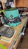 Photo of green fabric tool belt filled with hand tools on wooden surface, showing pliers, screwdrivers, and other tools, with black cushion and various items in background.