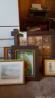 Photo showing multiple framed artworks leaning against wall with a wood paneled background. Includes a mix of landscape, lighthouse, and still life paintings.