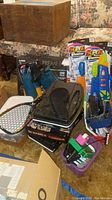 Photo showing boxed items including Scrabble games, Artskills artist case, water guns, tennis racket, and flippers on floral couch.