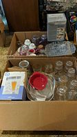 Overview of assorted glassware and serveware items packed in two boxes showing clear glass glasses, ceramic mugs, plastic mason jar style glasses, and covered casserole dishes.