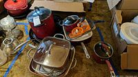 Overview of assortment of pots and pans, clear glass teapot, egg mold, and sharpener arranged on carpet.