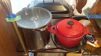 Photo showing Nesco roaster oven, stainless steel stock pot with glass lid, red enameled cast iron stock pot, and black vegetable grill pan on table.