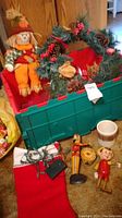 Green storage tub containing fals wreath and stuffed scarecrow along with holiday items visible on floor including ceramic crock, wooden puppets, and string lights.