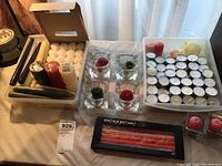 Various candles and holders arranged on a table including white tea lights, square glass holders containing colored votive candles, and more candles in a plastic container.