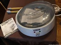 Full view of the Conair heating unit with clear lid, manual, and wooden tongs with blue tips on a table.