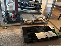 Photo showing MultiChef warming plate lying flat on carpet with paper instructions on top, and various ovenware and accessories on angled metal shelf above.