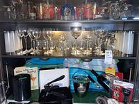 Wide view of glassware on a shelf including wine, cocktail, Irish coffee and shot glasses with other bar items beneath.