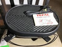 Black electric grill with ridged surface, power cord, and Tefal Gourmet Party Set manual on top.