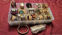 Overview showing about 21 ladies watches and metal bracelets in plastic container on red fabric surface.
