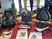 Photo showing the three kitchen appliances displayed with their respective instruction manuals on a red tablecloth.