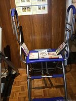 Front view of the Pilates Pro Chair with blue padding on handles and seat, showing the metal frame and resistance springs, with instructional books and papers on the seat.