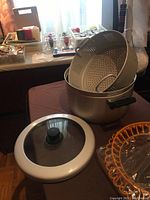 Five quart aluminum pasta pot with strainer insert, black handles, and clear glass lid with black knob. Orange plastic chip maker visible beside the pot.