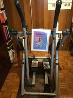 Full view of the Tony Little Gazelle Freestyle exercise machine in a room, showing foot pedals, handlebars with foam grips, and metal frame.