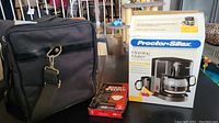 Photo showing black zippered travel bag, boxed Proctor Silex coffeemaker, and box of coffee filters on table.