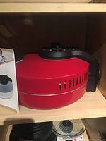 Red Wolfgang Puck pizza baker on a wood shelf, showing side venting and black handle.