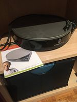 Wolfgang Puck branded electric crepe maker, round black unit with power cord seen coiled on side, resting on shelf with use and care instructions booklet.
