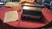 Front and top view of the Wolfgang Puck Tri Grill and Panini Press closed on a table with instruction sheet beside it, showing stainless steel finish and control knobs.