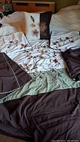 Comforter set laid out on bed showing embroidered floral design on cream background with brown and green fabric accents and matching pillow shams and throw pillow