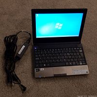 Front view of one Acer Aspire One netbook open, powered on displaying Windows 7 desktop, with power adapter cable beside it.