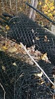 Rolls of black chain link fence and steel posts leaning against trees outdoors