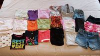 Photo of 18 assorted girls tops including t-shirts, long sleeve shirts, sweaters, and tank tops laid out on a bed.