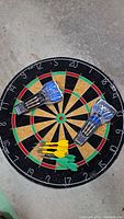 Full view of dart board with six plastic darts (three yellow, three green) arranged on it, and two sets of Eastpoint metal darts in plastic cases.