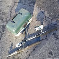 Two telescoping fishing rods with spinning reels laid on ground next to green plastic table box containing fishing gear.