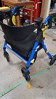 Three-quarter front view of blue rolling walker showing black padded seat, black fabric backrest, and four caster wheels on concrete floor.