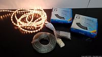 Four rolls of rope lights displayed with packaging boxes and accessories such as clips and zip ties on black background. One white rope light is plugged in and illuminated.