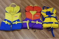 Three life jackets lying on a wooden floor—Roots size C yellow and blue life jacket, Coleman orange and purple size B life jacket, and yellow and blue Coleman size B life jacket.
