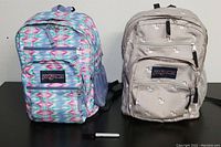 Front view of two Jansport backpacks: one colorful geometric pattern, one beige with swans.