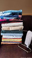 Stack of 7 bath towels in solid colors and 5 colorful beach towels folded on sofa with fingertip towel on metal stand beside them.