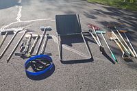 Overview photo showing wide variety of yard tools laid out on asphalt including rakes, shovels, brooms and a blue folding yard seat.