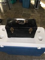 Front view showing the black hard suitcase with brass buckles and handle on top.