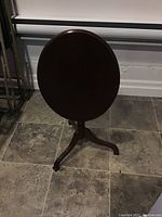 Front view of oval Bombay flip table with top in vertical position, showing dark wooden finish and tripod base.