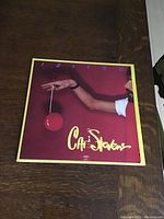 Front cover of Cat Stevens Izitso vinyl record showing person holding a red yo-yo on red background with yellow border and text