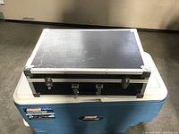 Photo of black hard carrying case resting on a blue cooler, showing top and front sides including handle and latches