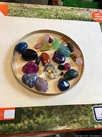 Round beige tray holding 21 assorted polished and rough mineral and gemstone specimens from Madagascar in multiple colors.