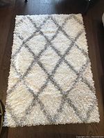 View of the entire cream-colored shag area rug with grey diamond lattice pattern on dark wood floor.