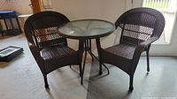 Full view of the outdoor bistro set showing round glass top table and two dark brown wicker-style chairs.